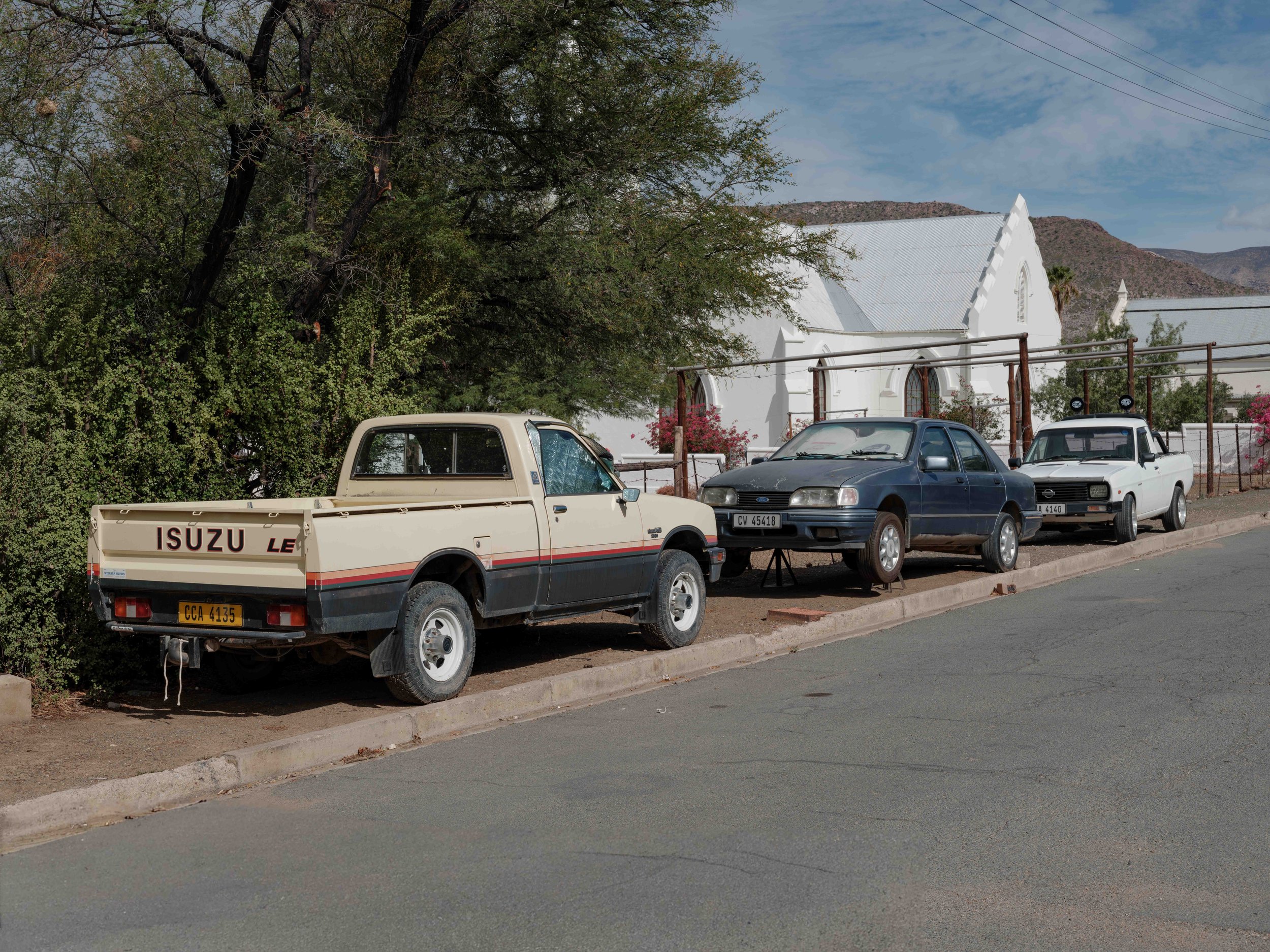Parked vehicles on a quiet street with a white building and hills beyond